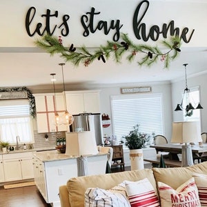 Puede incluir: Una sala de estar con un sof&aacute; de color beige y almohadas a rayas rojas y blancas. El sof&aacute; tiene una manta de cuadros rojos y negros. Las palabras "Let's stay home" est&aacute;n escritas en letras negras en la pared sobre el sof&aacute;. Hay una guirnalda de vegetaci&oacute;n con bayas rojas colgando sobre las palabras.