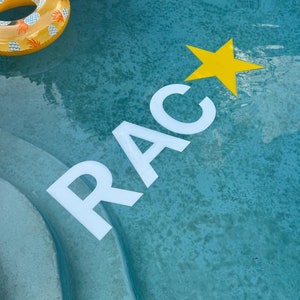 May include: White floating letters spelling RAC and a yellow floating star in a blue swimming pool. A yellow and white pineapple patterned inflatable ring floats in the upper left corner, partially visible above the water surface.