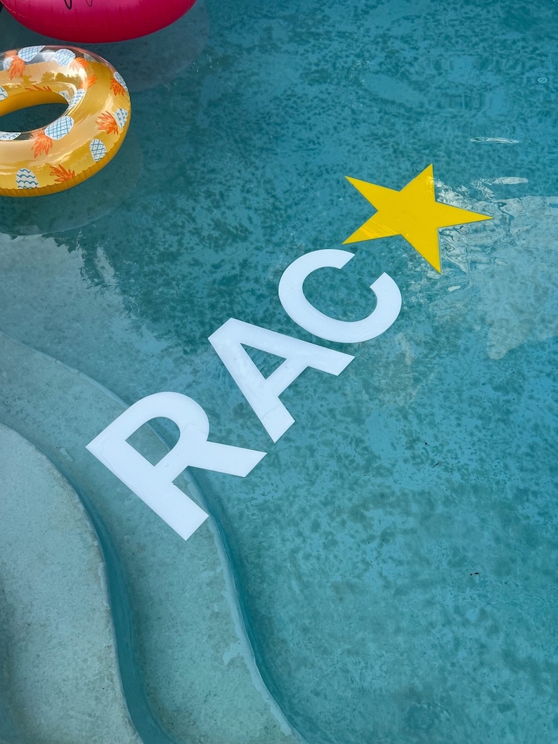 May include: White floating letters spelling RAC and a yellow floating star in a blue swimming pool. A yellow and white pineapple patterned inflatable ring floats in the upper left corner, partially visible above the water surface.