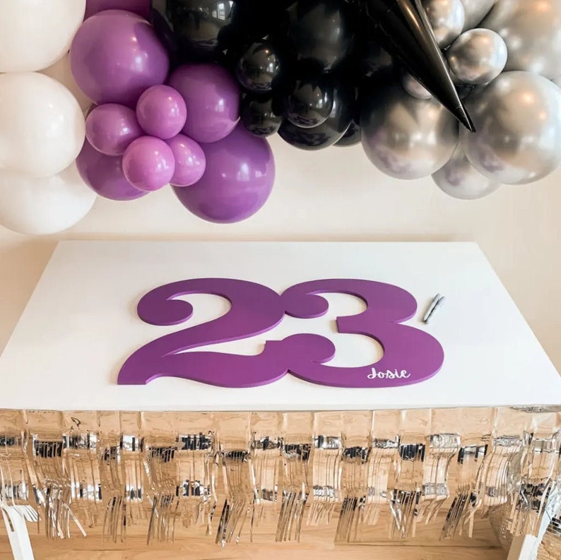 May include: Purple number 23 sign with the name Josie written below. The sign is on a white table with a gold fringe tablecloth. The background is a wall with a purple, black, and silver balloon garland.
