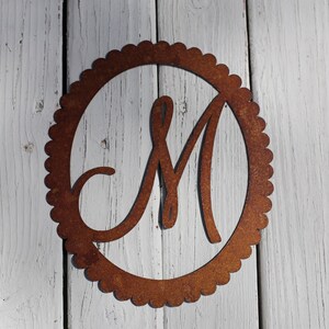 May include: A rusty metal monogram with the letter "M" in a decorative script font. The letter is centered in a circle with a scalloped edge.