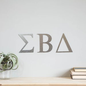 May include: Silver metal letters spelling out "ΣΒΔ" on a white wall. The letters are mounted on the wall and are a popular choice for sorority houses or rooms.