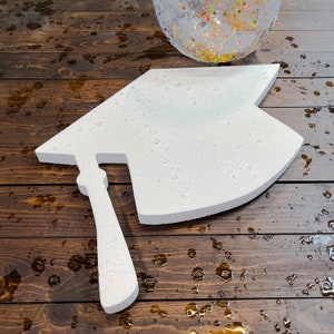 May include: White foam graduation cap cutout with a raised tassel. The cap is on a brown wooden surface with water droplets.