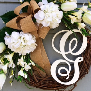 May include: A wreath made of grapevine with a burlap bow and white flowers. The wreath has a white wooden letter "E" in the center.