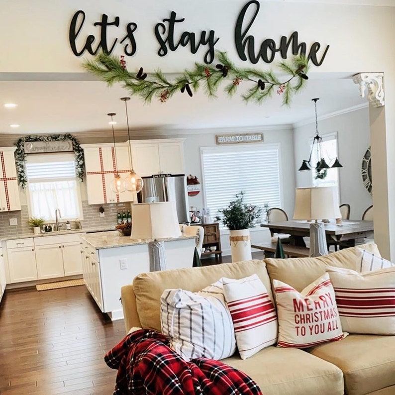 Puede incluir: Una sala de estar con un sof&aacute; de color beige y almohadas a rayas rojas y blancas. El sof&aacute; tiene una manta de cuadros rojos y negros. Las palabras "Let's stay home" est&aacute;n escritas en letras negras en la pared sobre el sof&aacute;. Hay una guirnalda de vegetaci&oacute;n con bayas rojas colgando sobre las palabras.