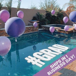 May include: White foam letters spelling "#GRAD" floating in a swimming pool with purple balloons. The text "SHIPS IN 1 BUSINESS DAY!" is on a purple background.