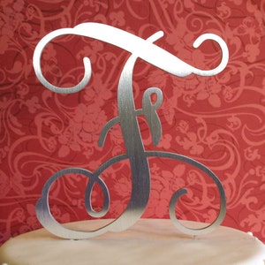 May include: Silver metal letter F cake topper with a decorative script font. The topper is on a white cake with a red floral pattern background.