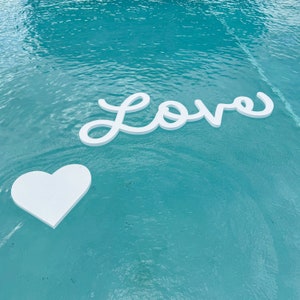 May include: White cutout letters spelling "LOVE" floating in a turquoise blue pool of water. A white heart cutout is floating below the word "LOVE".