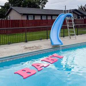 May include: A backyard swimming pool with a blue slide and a pink foam word "BABY" floating in the water. The pool has a blue tile border and is surrounded by a concrete deck, a red fence, and green grass.
