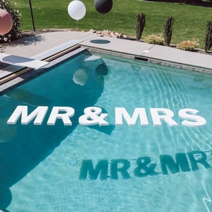 May include: White letters spelling "MR & MRS" float in a blue swimming pool. The letters are reflected in the water.
