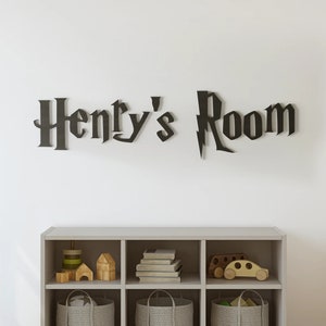 May include: A white wall with a black wooden sign that reads "Henry's Room" in a Harry Potter-inspired font. Below the sign is a gray wooden shelf with six cubbies. The cubbies are filled with toys, including a wooden car, a stack of books, and a basket.