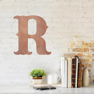May include: A large, rusty brown metal letter R with a decorative font. The letter is mounted on a white brick wall. There is a small potted plant, a white mug, a stack of books, and a vase with dried flowers on a white countertop in front of the wall.