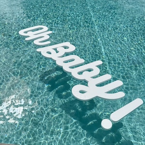 May include: White foam letters spelling "Oh Baby!" floating in a turquoise swimming pool.