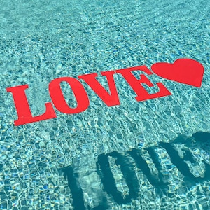 May include: Bright red, flat, cutout letters spelling the word LOVE followed by a red heart shape, floating on the surface of clear, rippling blue swimming pool water, with a dark shadow of the letters cast onto the pool floor below.