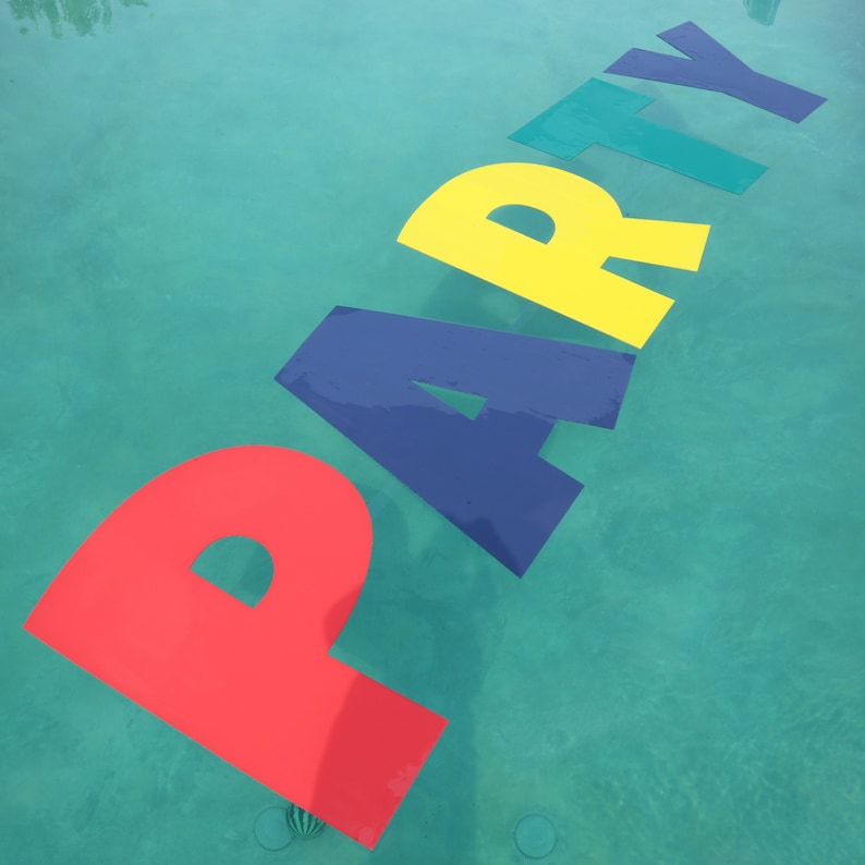 May include: A set of large, colorful floating letters spelling the word PARTY on the surface of a swimming pool. The letter P is red, the A is dark blue, the R is yellow, the T is teal, and the Y is dark blue, all resting on turquoise water.