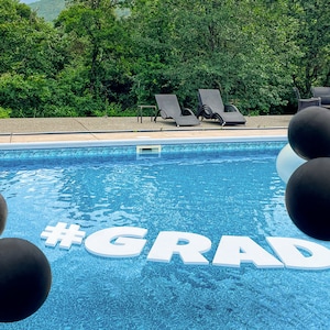 May include: A swimming pool with black and white balloons floating in the water. The words "#GRAD" are spelled out in large white letters on the surface of the water.