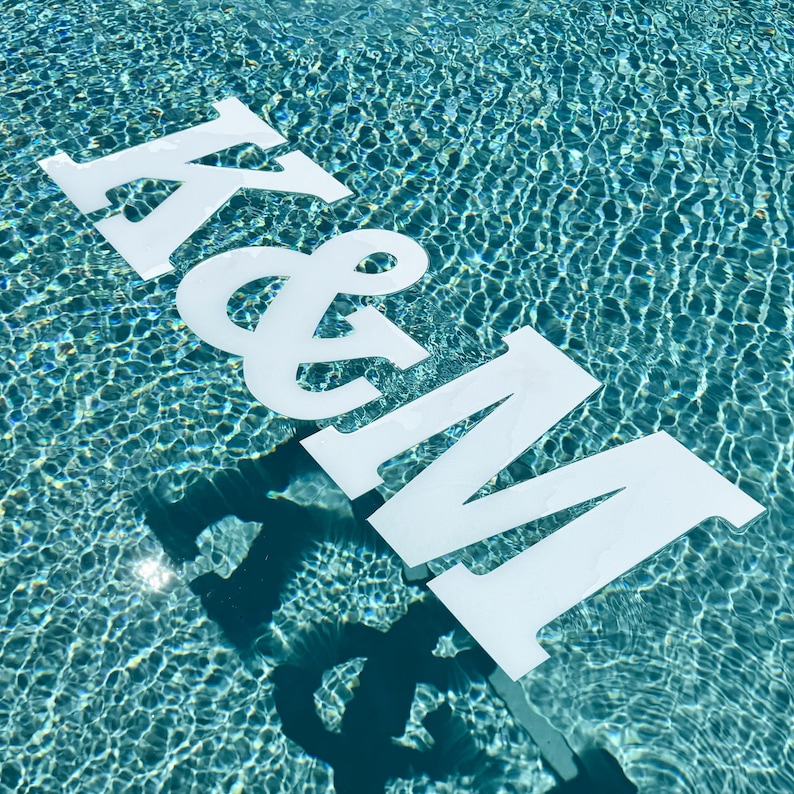 May include: Large white capital letters K and M connected by an ampersand symbol floating on the surface of a blue swimming pool. The letters are flat and opaque, casting dark shadows onto the rippling water beneath them in the bright sunlight.