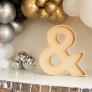 May include: A large, beige, wooden ampersand sign with a decorative, cutout design. The sign is sitting on a white surface with silver and gold balloons and disco balls in the background.