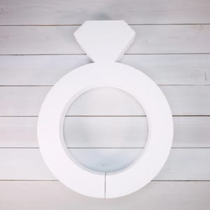 May include: White foam ring with a diamond-shaped top, resembling a large engagement ring. The ring is on a white wood background.
