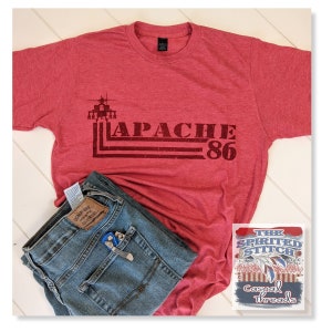 May include: Red heather t-shirt with a distressed graphic of a helicopter and the text "APACHE 86". The t-shirt is paired with blue jeans and a small multi-tool.