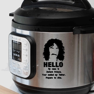 May include: A stainless steel Instant Pot with a black lid and handle. The pot has a digital display and control panel. A black graphic of a face with the text "HELLO My name is Instant Potoya. Your sealed my father. Prepare to dine." is on the side.