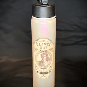 May include: A pearlescent water bottle with a black lid and handle. The bottle features a vintage-style label with the words "Elixir of Life" and a graphic of a person. The label also includes the text "Extra Quality" and "Shake Before Use."