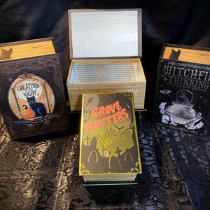 May include: Three decorative book-shaped boxes. The left box features a black cat and pumpkin with the text 'Creature of the Night'. The right box shows a cauldron with the text 'Witchful Thinking'. The center box is open, displaying a 'Grave Matters' cover.