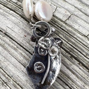 K&ouml;nnte beinhalten: Eine silberne Anh&auml;ngerkette mit gro&szlig;en, unregelm&auml;&szlig;igen wei&szlig;en Perlen. Der Anh&auml;nger hat ein strukturiertes, abstraktes Design mit floralem Motiv. Die Halskette wird auf einer verwitterten Holzoberfl&auml;che pr&auml;sentiert.
