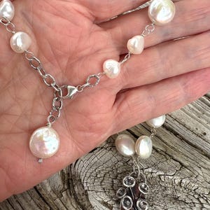 K&ouml;nnte beinhalten: Eine silberne Halskette mit einer Kette aus wei&szlig;en, unregelm&auml;&szlig;ig geformten Perlen. Die Halskette hat einen dekorativen Silberanh&auml;nger mit floralem Design. Der Verschluss ist ebenfalls silberfarben.