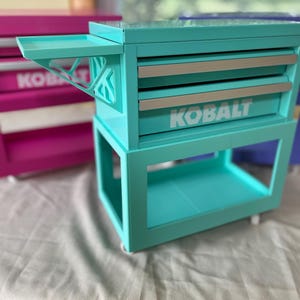 May include: A bright turquoise metal tool chest with two drawers, each featuring a silver handle and the white text KOBALT printed on the front. The unit includes a side shelf with a geometric support bracket and sits on small wheels.