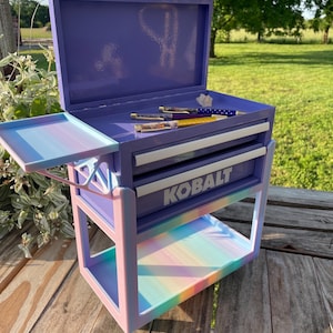 May include: A purple metal Kobalt tool chest with two drawers and an open top lid. The unit features a multi-colored pastel gradient frame and a side tray. Two pens with patterned barrels rest on the top surface inside the open chest.
