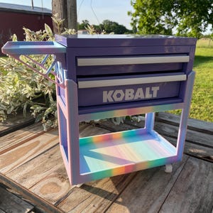 Könnte beinhalten: Ein lila und pastellfarbener Werkzeugwagen mit dem Wort "KOBALT" auf der Vorderseite. Der Wagen hat zwei Schubladen und ein unteres Regal. Der Wagen ist auf Rädern und hat ein ausziehbares Tablett.