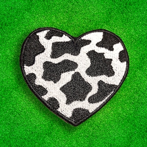 May include: A black and white cow print heart-shaped patch. The patch is made of fabric and has a stitched edge.