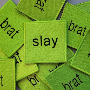 May include: A close-up of a stack of green felt patches with the words "slay", "brat", and "br" embroidered on them.