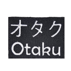 May include: Black rectangular patch with white embroidered text. The top line features Japanese characters, and the bottom line reads "Otaku" in white, sans-serif font. The patch is likely for clothing or accessories.