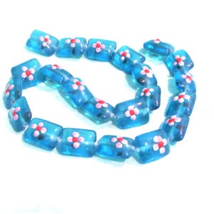 Könnte beinhalten: Eine Kette aus durchscheinenden, blauen, rechteckigen Perlen. Jede Perle hat eine kleine, weiße Blume mit roter Mitte. Die Perlen sind aufgefädelt und bilden eine flexible, gebogene Form vor weißem Hintergrund. Diese Perlen sind wahrscheinlich für die Schmuckherstellung.