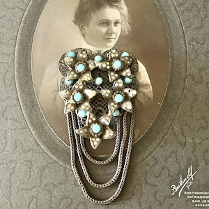May include: A silver and turquoise floral brooch with a long chain pendant. The brooch is in the shape of a heart and features several turquoise stones set in silver.
