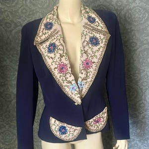 May include: A navy blue blazer featuring a decorative floral pattern on the lapels and pockets. The floral design incorporates pink, blue, and green details against a cream backdrop. The blazer fastens with a single button.