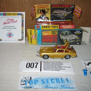 May include: A gold-colored Corgi Toys model of James Bond's Aston Martin DBS, with the number 261 on the box. The image includes the car, boxes, a certificate, and other accessories. The text "007" is visible.