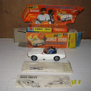 May include: A white Corgi Toys Toyota 2000 GT model car from the James Bond film, with a red interior and figures inside. The packaging includes the original box and instructions. The box features images of the car and James Bond.
