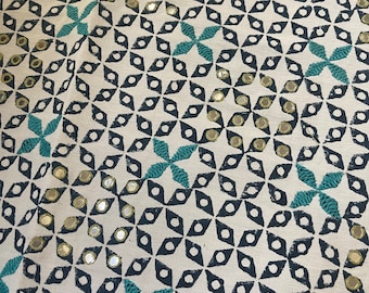 May include: Cream-colored fabric with a repeating pattern of navy blue diamond shapes, teal floral accents, and small, round mirrored embellishments. The design creates a geometric and textured appearance, suitable for home decor or textile projects.