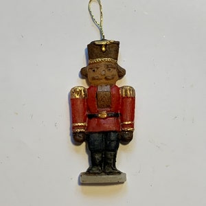 Puede incluir: Un peque&ntilde;o adorno de cascanueces rojo y dorado con un sombrero marr&oacute;n y una base blanca. El cascanueces sostiene una peque&ntilde;a espada dorada.