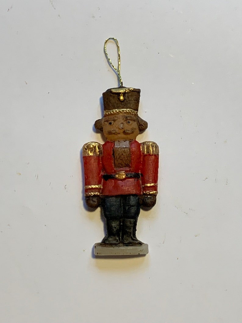 Puede incluir: Un peque&ntilde;o adorno de cascanueces rojo y dorado con un sombrero marr&oacute;n y una base blanca. El cascanueces sostiene una peque&ntilde;a espada dorada.