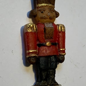 Puede incluir: Un adorno de cascanueces vintage hecho de madera pintada. El cascanueces lleva un uniforme rojo y dorado con un sombrero negro y una base blanca.