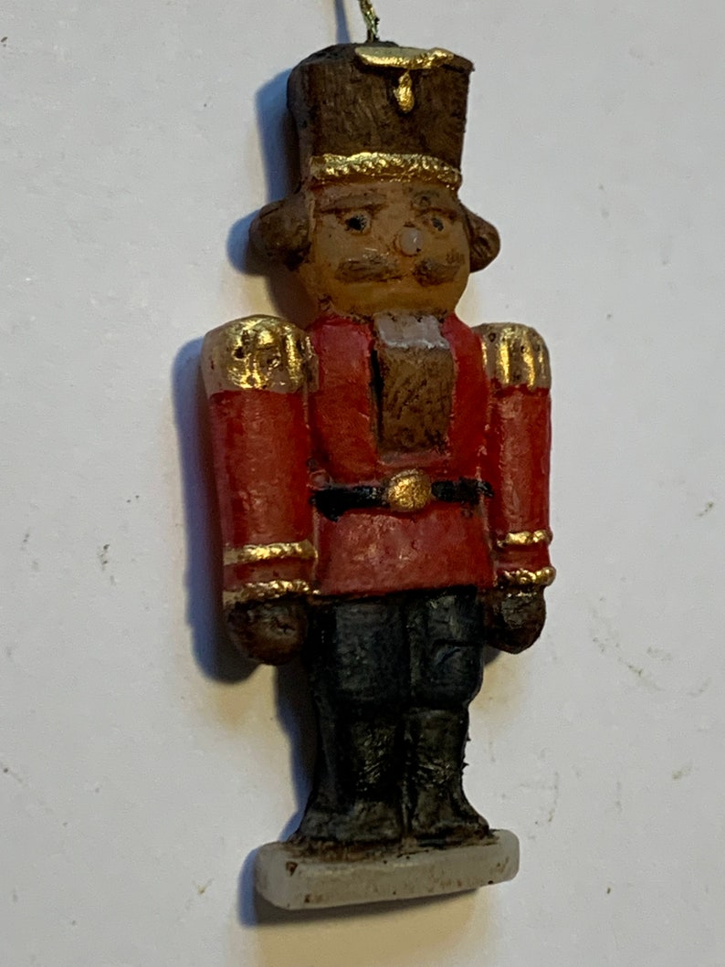 Puede incluir: Un adorno de cascanueces vintage hecho de madera pintada. El cascanueces lleva un uniforme rojo y dorado con un sombrero negro y una base blanca.