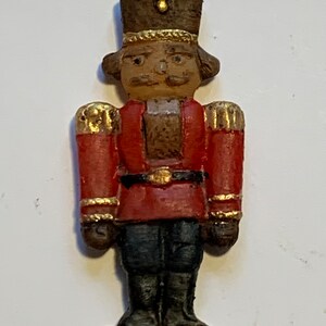 Puede incluir: Un adorno de cascanueces de estilo vintage con un uniforme rojo y dorado, un sombrero marr&oacute;n y una cara pintada. El adorno est&aacute; hecho de cer&aacute;mica y tiene una base blanca.