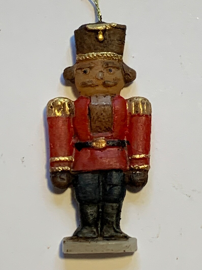 Puede incluir: Un adorno de cascanueces de estilo vintage con un uniforme rojo y dorado, un sombrero marr&oacute;n y una cara pintada. El adorno est&aacute; hecho de cer&aacute;mica y tiene una base blanca.