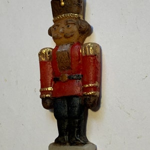 Puede incluir: Un adorno de cascanueces de madera vintage con un uniforme rojo y dorado, un sombrero marr&oacute;n y una cara pintada. El adorno tiene una cuerda dorada para colgar.