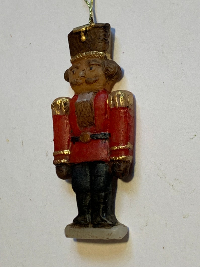 Puede incluir: Un adorno de cascanueces de madera vintage con un uniforme rojo y dorado, un sombrero marr&oacute;n y una cara pintada. El adorno tiene una cuerda dorada para colgar.
