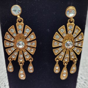 May include: Gold tone dangle earrings with a large crystal center surrounded by smaller crystals. The earrings have a teardrop shape with three smaller teardrop crystals hanging from the bottom.